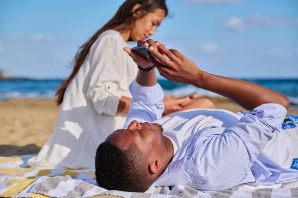 Νεαρό πολυπολιτισμικό ζευγάρι που βρίσκεται στην παραλία χρησιμοποιώντας smartphones — Φωτογραφία Αρχείου