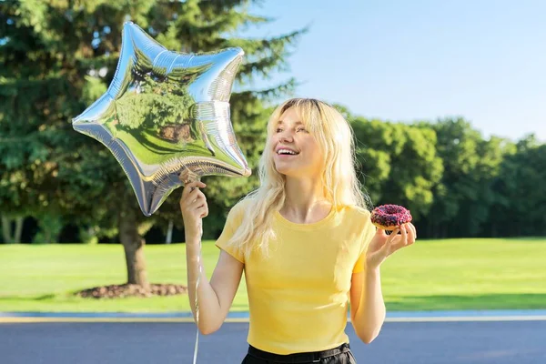 Retrato de hermosa rubia adolescente con rosquilla de pastel y globo de plata en el parque —  Fotos de Stock