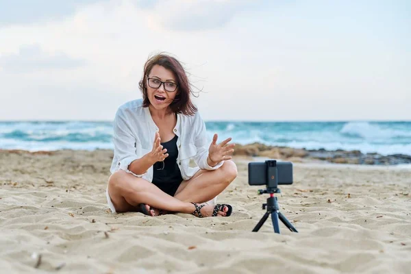 Жінка середнього віку сидить на пляжі зі смартфоном за допомогою відеодзвінка — стокове фото