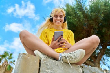 Kulaklıklı genç kız akıllı telefona bakıyor.