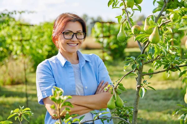 Potret wanita paruh baya yang tersenyum dan percaya diri dengan lengan bersilang di kebun buah, dekat pohon pir — Stok Foto