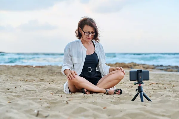 Mujer de mediana edad sentada en la playa con smartphone usando videollamada — Foto de Stock