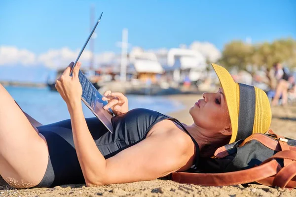 Mature woman sunbathing on the beach in hat with laptop — стоковое фото