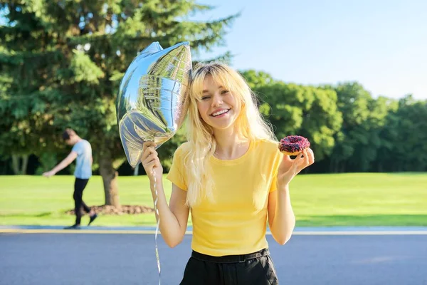 Retrato de hermosa rubia adolescente con rosquilla de pastel y globo de plata en el parque —  Fotos de Stock