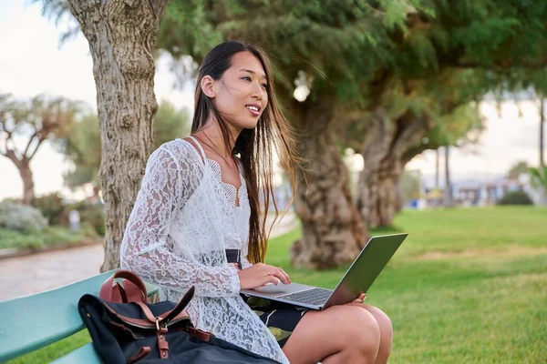 Όμορφη νεαρή Ασιάτισσα γυναίκα που χρησιμοποιεί φορητό υπολογιστή κάθεται σε ένα υπαίθριο πάγκο — Φωτογραφία Αρχείου