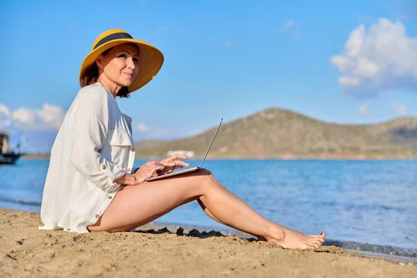 Mujer madura en sombrero usando el ordenador portátil en la playa del mar — Foto de Stock