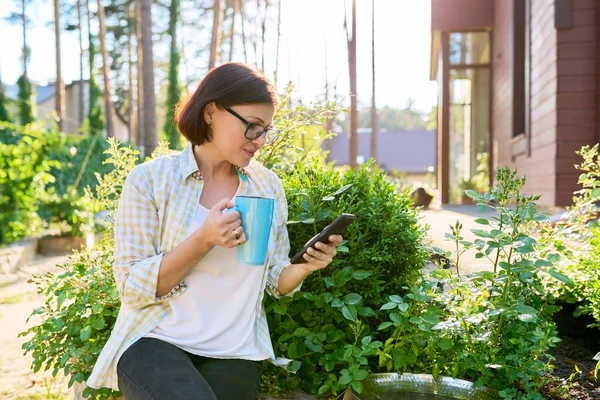 Μεσήλικη γυναίκα χαλαρώνει στον κήπο με smartphone και κύπελλο στα χέρια της — Φωτογραφία Αρχείου