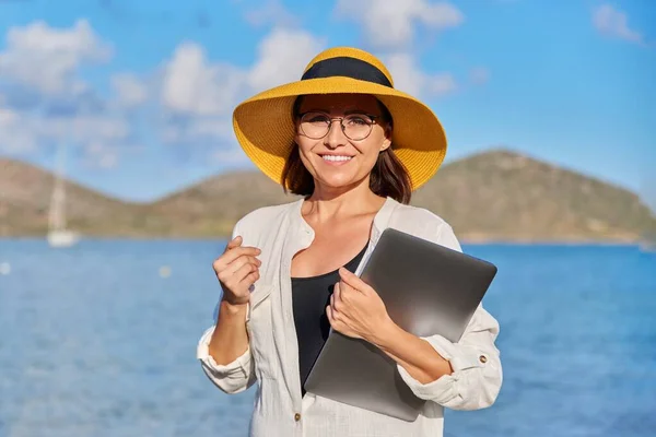 지푸라기 모자를 쓰고 해변에 랩탑을 들고 있는 행복 한 성인 여성의 모습 — 스톡 사진