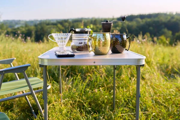 Kubki, szlifierka, ziarna kawy na składanym stole do kempingu na świeżym powietrzu — Zdjęcie stockowe