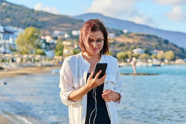스마트폰으로 헤드폰을 끼고 해변에서 쉬고 있는 중년 여성 — 스톡 사진