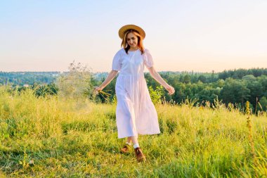 Hasır şapkalı, beyaz elbiseli romantik kadın günbatımının tadını çıkarıyor.