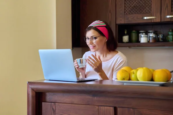 Mujer de mediana edad bebiendo café hablando en videollamada usando computadora portátil, cocina en casa — Foto de Stock