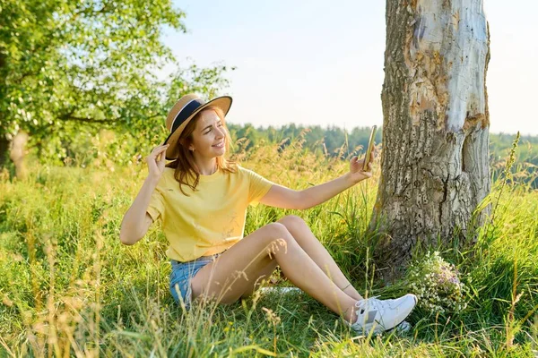 Vuxen kvinna i halm hatt avkopplande, sitter på gräs, tittar i smartphone webbkamera, gör videosamtal — Stockfoto