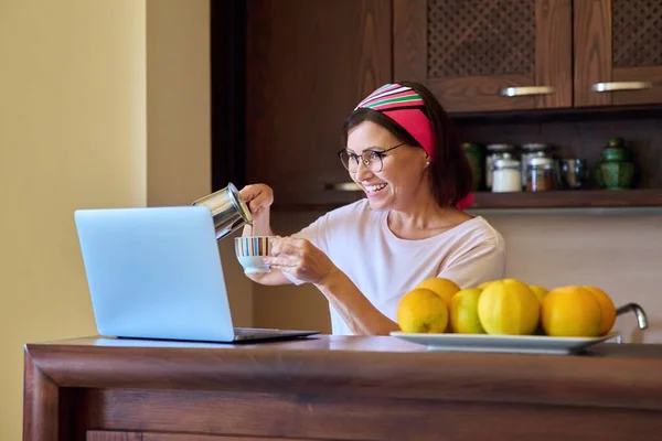 Mujer de mediana edad bebiendo café hablando en videollamada usando computadora portátil, cocina en casa — Foto de Stock