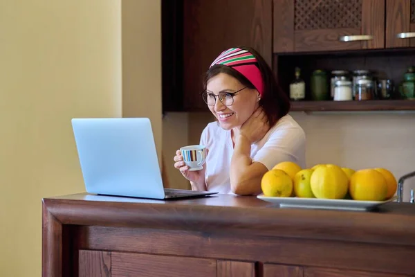 Mujeres de 40 años de edad descansando en casa con una taza de café mirando a la computadora portátil — Foto de Stock