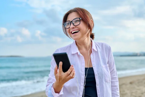 Ευτυχισμένη μεσήλικη γυναίκα στη θάλασσα διακοπές απολαμβάνοντας ακουστικά με smartphone — Φωτογραφία Αρχείου