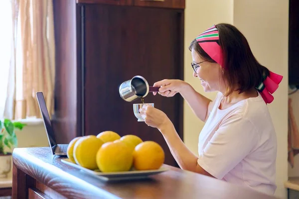 Жінка середнього віку п'є каву, розмовляючи на відео виклик за допомогою ноутбука, кухні вдома — стокове фото