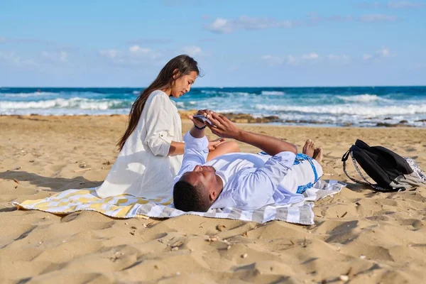 年轻夫妇在海滩上放松，坐在沙滩上，拿着智能手机 — 图库照片