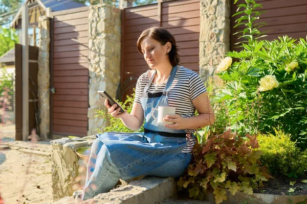 Μεσήλικη γυναίκα χαλαρώνει στον κήπο με smartphone και κύπελλο στα χέρια της — Φωτογραφία Αρχείου