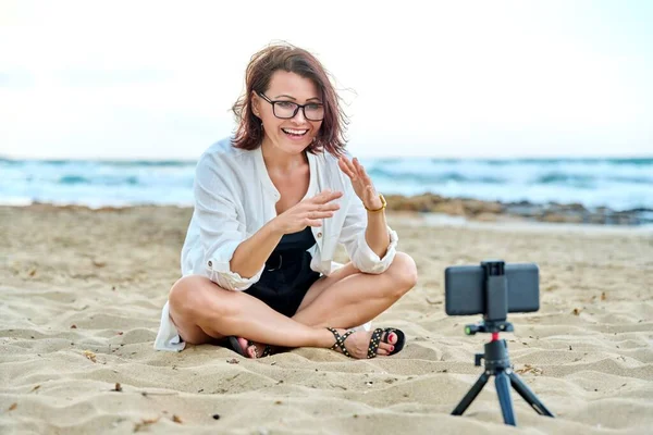 해변에 앉아 스마트폰으로 비디오 통화를 하는 중년 여성 — 스톡 사진