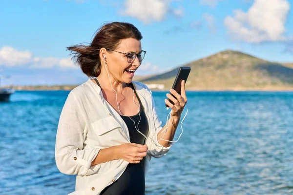 Ευτυχισμένη 40χρονη γυναίκα με ακουστικά και smartphone στην παραλία — Φωτογραφία Αρχείου