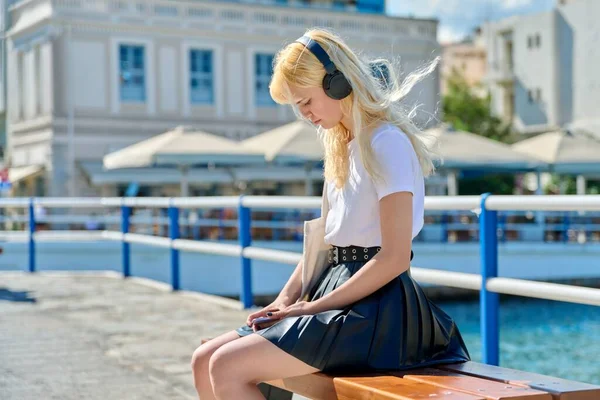 Сумна жінка-підліток в навушниках зі смартфоном, що сидить на морській набережній — стокове фото