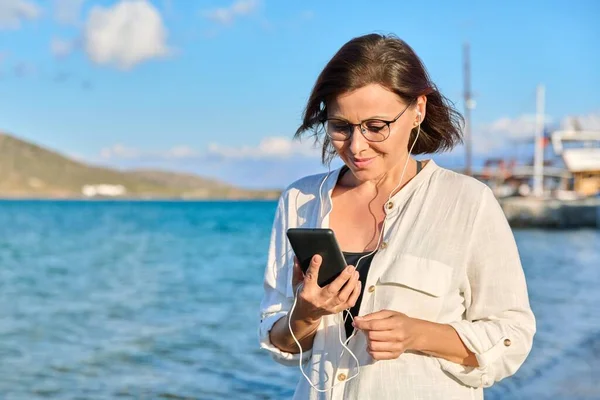 헤드폰을 끼고 해변에 스마트폰을 소지 한 행복 한 40 대 여성 — 스톡 사진