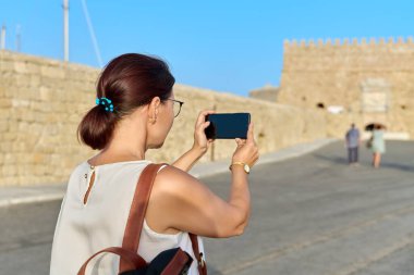 Akıllı telefondan fotoğraf çeken bir kadın turist, deniz kıyısındaki tarihi bir kale. Turizm, seyahat, insanlar, Yunanistan Girit Heraklion
