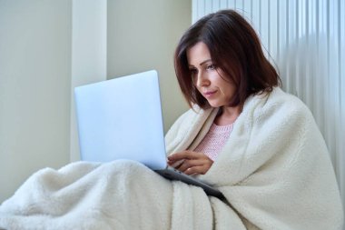 Evde ısıtma kaloriferinin yanında oturan kürklü bir kadın dizüstü bilgisayar kullanarak ısınıyor. Soğuk mevsim sonbahar kışı, yaşam tarzı, insanlar konsepti