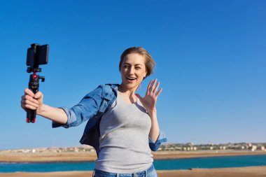 Orta yaşlı bir kadın deniz kıyısında açık havada akıllı telefondan video kaydediyor.