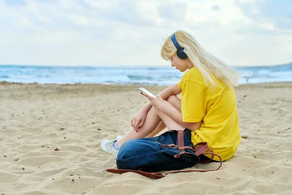 Mulher adolescente com mochila em fones de ouvido usando smartphone, na praia arenosa — Fotografia de Stock