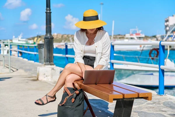 На відкритому повітрі, жінка середнього віку в солом'яному капелюсі сидить на лавці за допомогою ноутбука — стокове фото