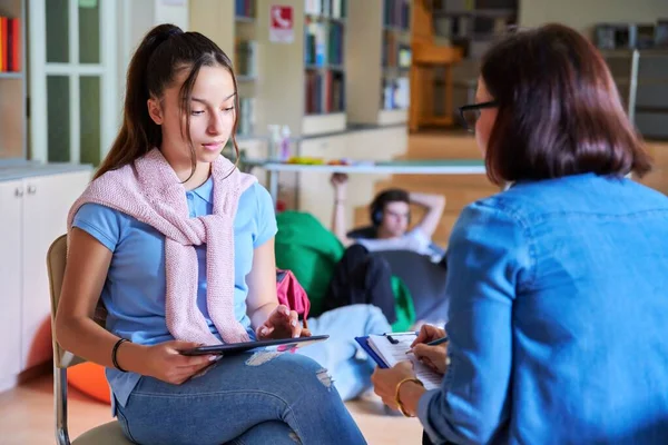 Teenage girl student talking with teacher mentor in the library.