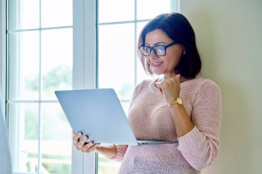 Pencerenin yanında dizüstü bilgisayar kullanan orta yaşlı bir kadın..