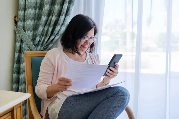 Medelålders överraskad kvinna med smartphone företagspapper sitter hemma — Stockfoto