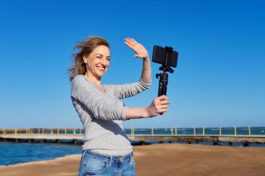 Orta yaşlı bir kadın deniz kıyısında açık havada akıllı telefondan video kaydediyor.