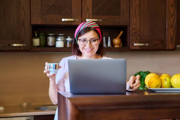 Medelålders kvinna i hemmet kök avkopplande med laptop för fritid och underhållning — Stockfoto