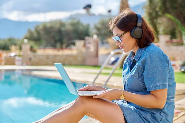 Donna di mezza età in cuffia utilizzando il computer portatile mentre seduto vicino alla piscina all'aperto — Foto Stock