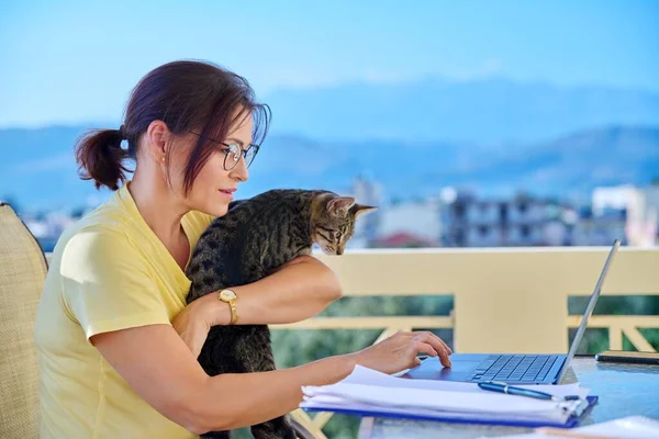 Жінка середнього віку працює в домашньому офісі на терасі з домашнім котом на руках — стокове фото