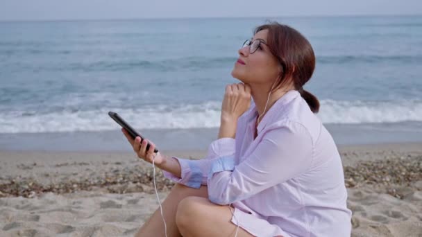 Donna di mezza età che si rilassa sulla spiaggia in cuffia con smartphone — Video Stock