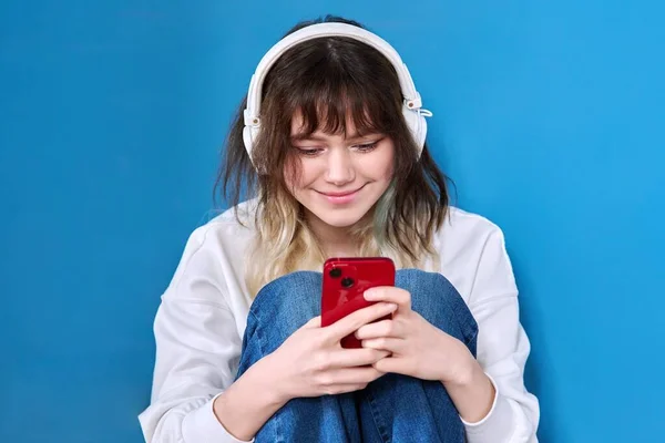 Teenage female in headphones with smartphone sitting on blue — Photo