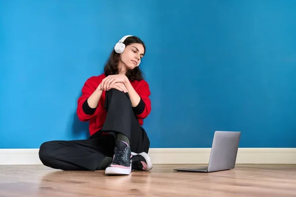 Jovem adolescente em fones de ouvido com laptop sentado no chão no fundo azul — Fotografia de Stock