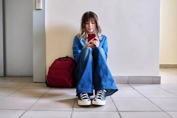 Студент-підліток в навушниках з рюкзаком смартфон, що сидить на підлозі біля стіни . — стокове фото