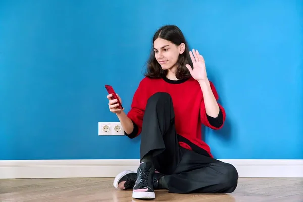 Teenage samec s chytrým telefonem sedí na podlaze na modrém pozadí — Stock fotografie