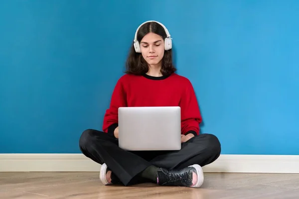Молодий хлопець-підліток в навушниках з ноутбуком, що сидить на підлозі на синьому фоні — стокове фото