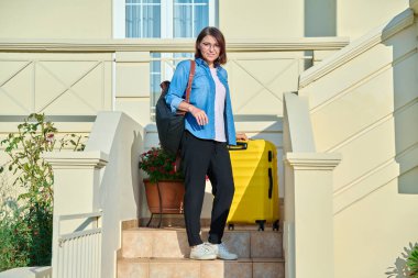 Evin merdivenlerinde çantalı orta yaşlı bir kadın var.