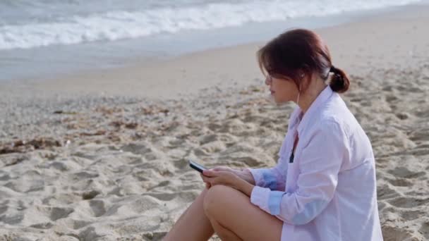 Donna di mezza età che si rilassa sulla spiaggia in cuffia con smartphone — Video Stock