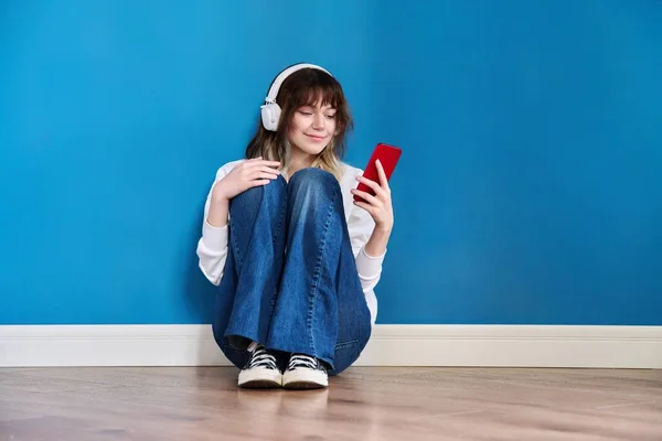 Teenage female in headphones with smartphone sitting on the floor on blue — Photo