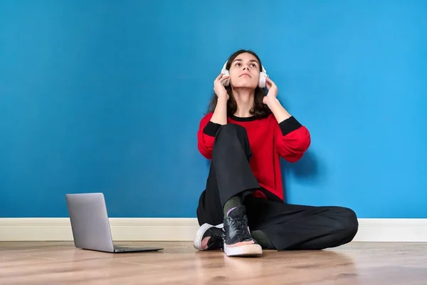 Joven adolescente en auriculares con portátil sentado en el suelo sobre fondo azul —  Fotos de Stock