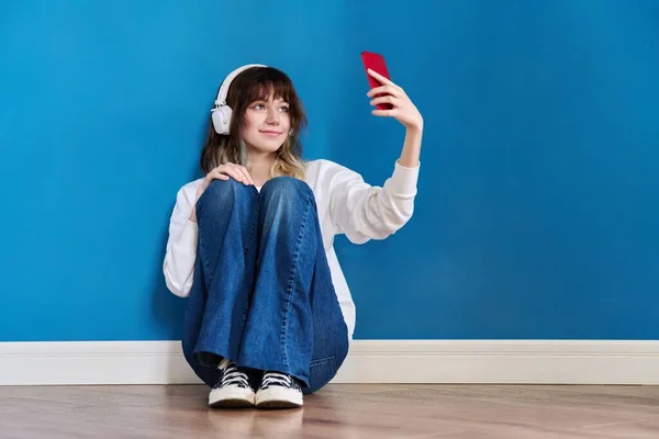 Dospívající žena ve sluchátkách s chytrým telefonem sedí na podlaze na modré — Stock fotografie